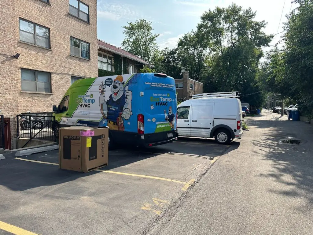 An unopened air conditioner in a box on the floor next to 2 vans.