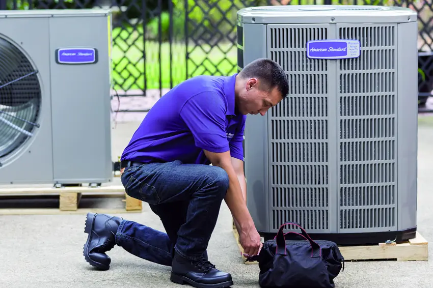 A man wearing a blue shirt kneeling down closing his duffel bag next to 2 american standard branded air conditioner systems.