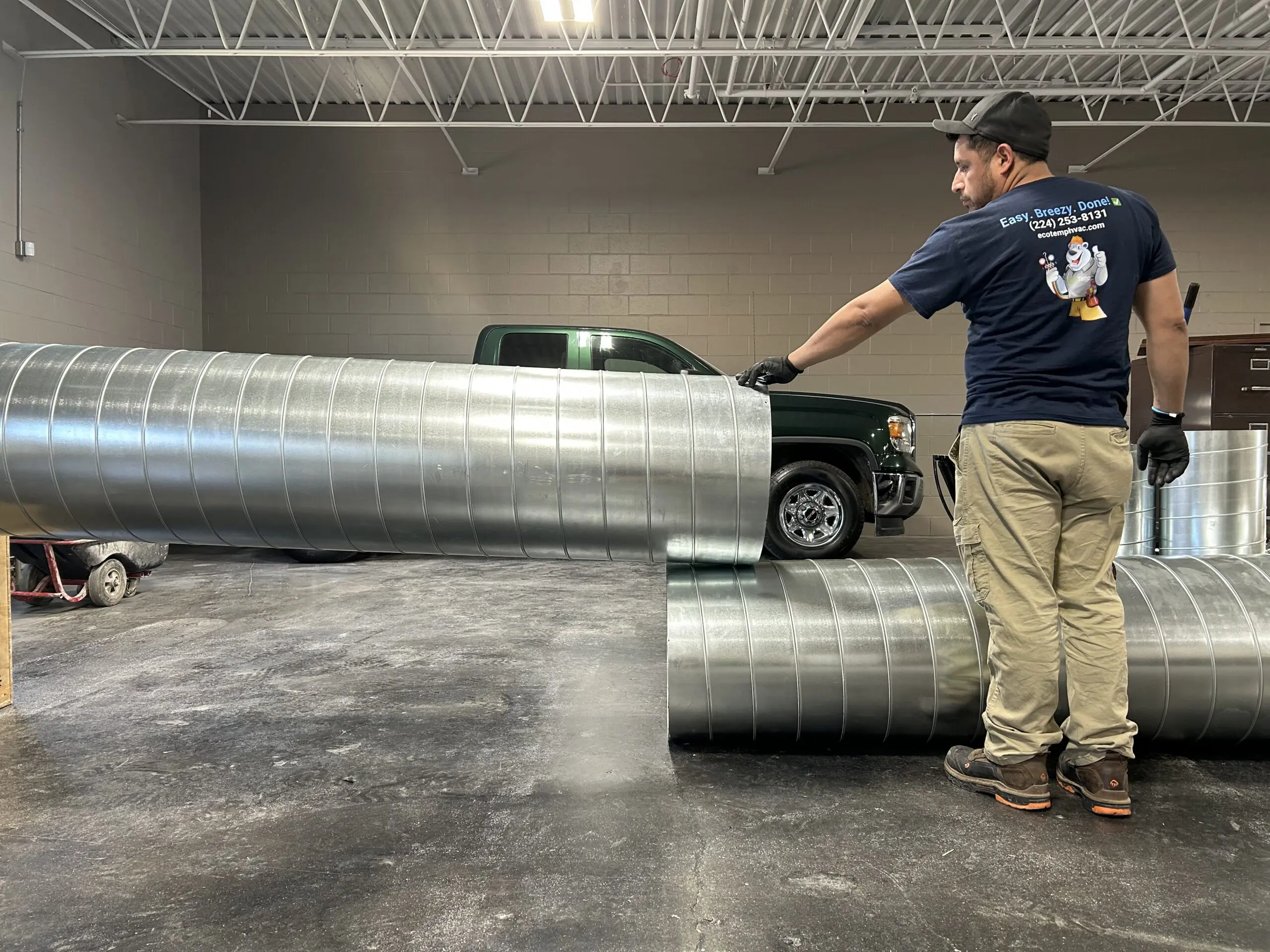 An HVAC technician standing in a well lit garage holding a piece of duct work with another air duct on the ground.