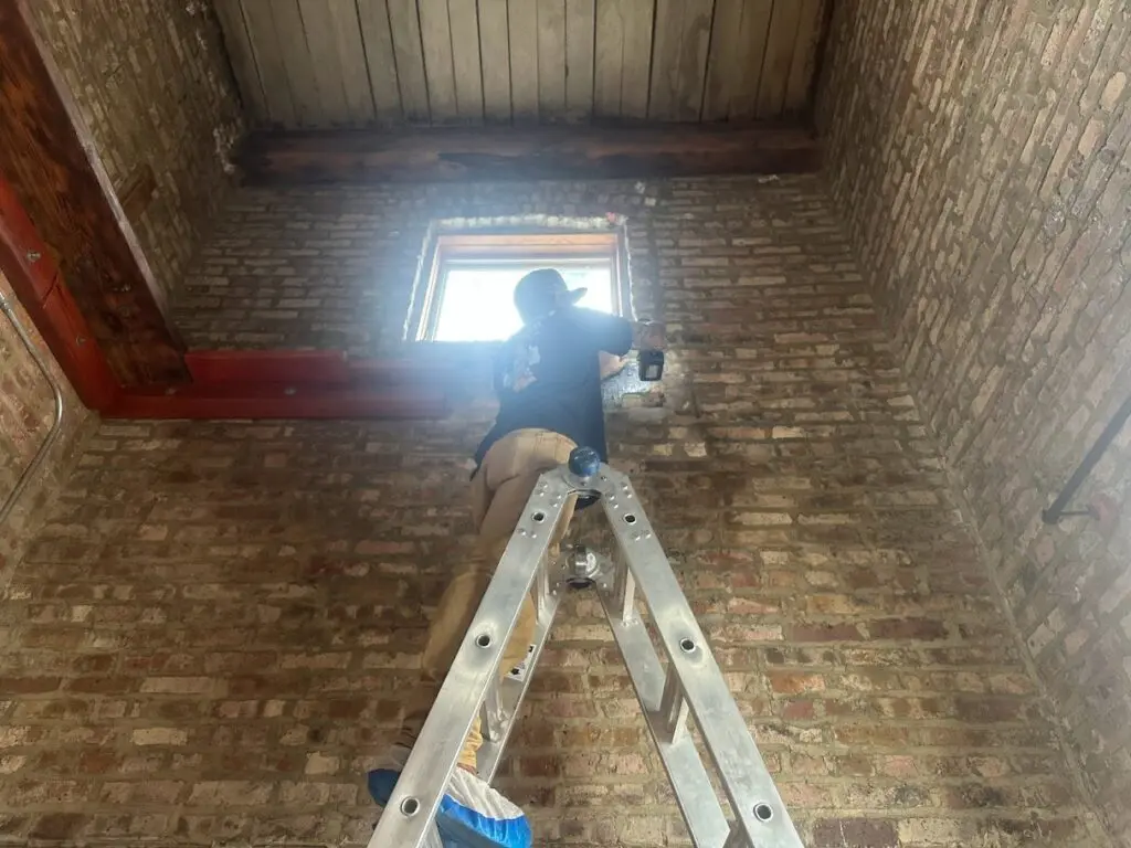 A HVAC technician standing on a ladder holding an electric drill.